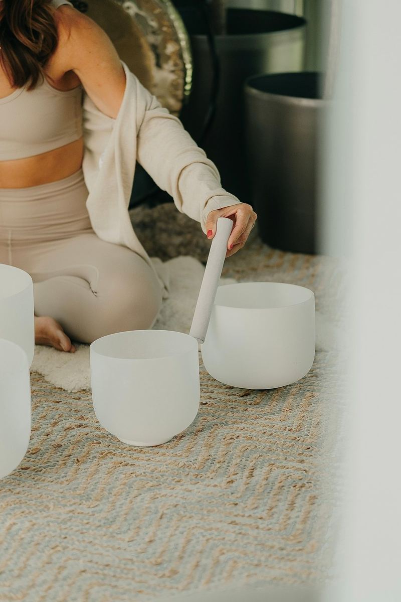 A woman in cream kneels behind white crystal singing bowls with a mallet.