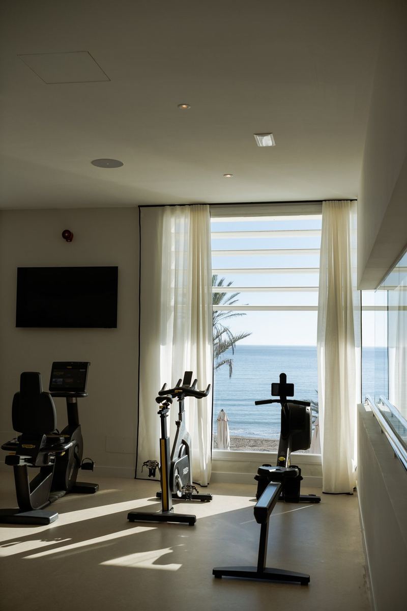 A bright hotel gym, a bike and a rower in front of white curtains and the sea.