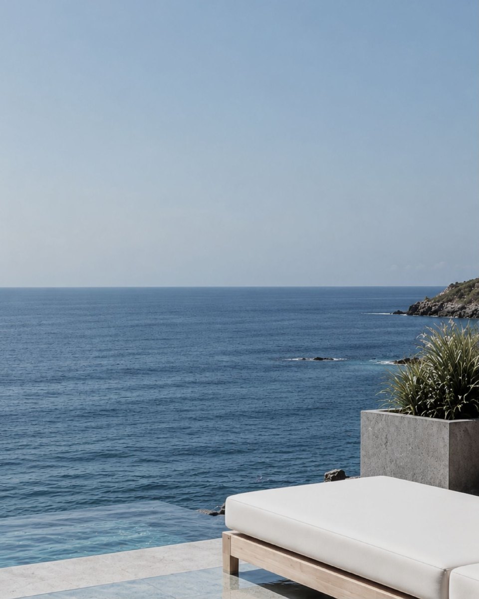 A sun lounger on a stone terrace by an infinity edge, the open sea and a rocky point beyond.