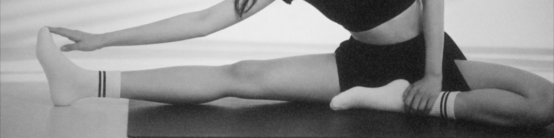 In black and white, a woman in a wide seated stretch on a mat, white crew grip socks with two dark stripes.